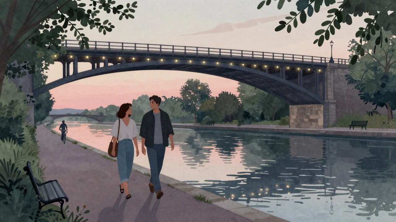 Two people walking along a canal at sunset, passing under railway arches with soft lights glowing, surrounded by peaceful greenery.